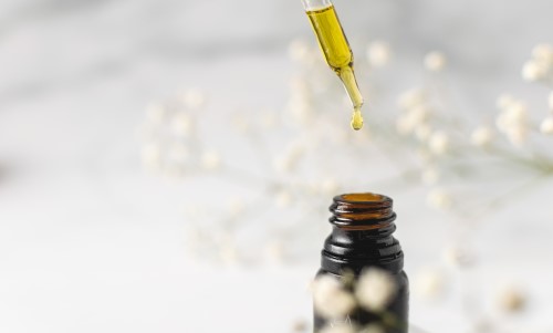 A close up view of a glass dropper filled with yellow liquid suspended over an amber colored bottle, dripping the liquid into the bottle.