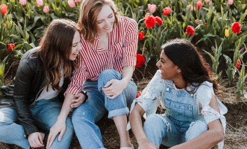 A group of female friends sit on the ground outside wearing jeans, long sleeved tops, while smiling and talking on a sunny day.